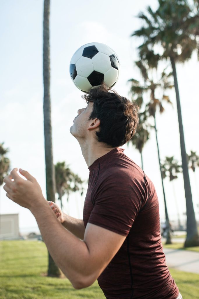 A person balances a soccer ball on their forehead while standing in an outdoor area with palm trees. The individual wears a maroon shirt and has arms raised, focusing on the ball. The sky is clear, and the background features grass and a few structures.