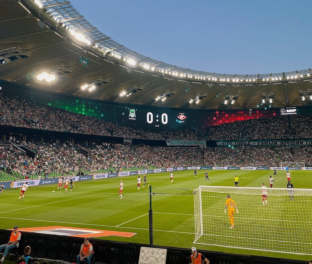 A wide-angle view of a soccer match in a large stadium packed with spectators. The scoreboard shows a 0-0 scoreline. Players are actively engaged on the field, with one team in red and white and the other in black and white. Bright stadium lights illuminate the scene.