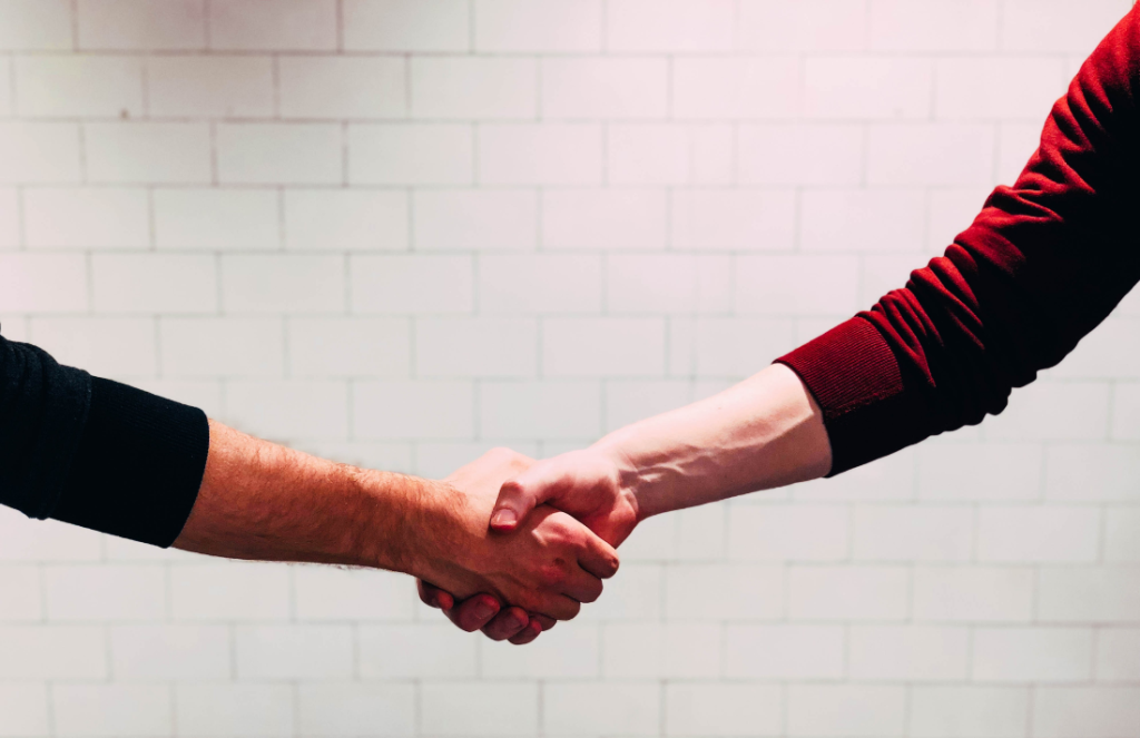 Two people shaking hands against a white tiled background. The person on the left is wearing a black sweater and the person on the right is wearing a red sweater. Only their extended arms and hands are visible in the image.