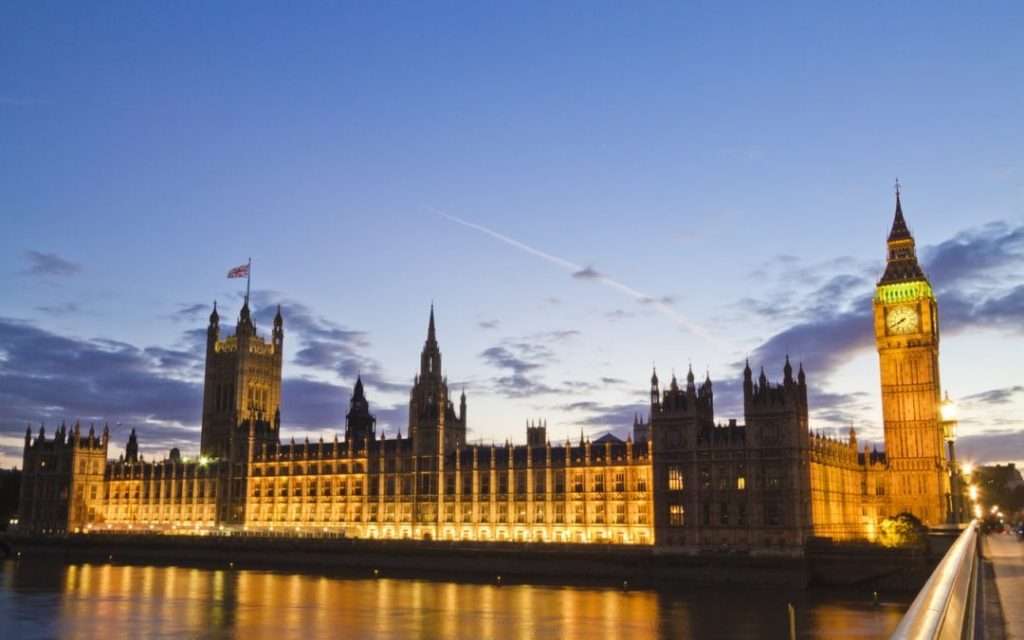 A nighttime view of the Houses of Parliament and Big Ben in London, illuminated by lights, with the River Thames in the foreground. The sky has a gradient from blue to twilight with some scattered clouds. The Union Jack flag is flying atop the building.