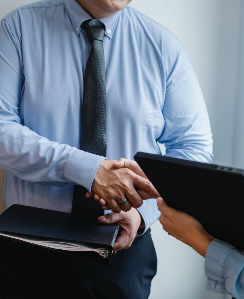 Two individuals in business attire are shaking hands. One person, in a light blue shirt and dark tie, uses both hands—one to grip a binder, the other to shake hands. The other person's hand and part of a tablet are visible, suggesting a professional setting.