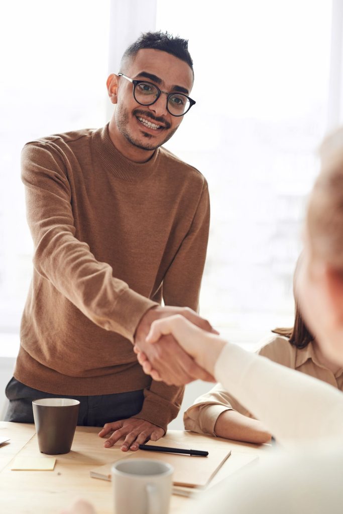 A man with glasses and a brown sweater is standing and shaking hands with someone seated, whose back is to the camera. They are in an indoor setting with mugs and documents on the table.