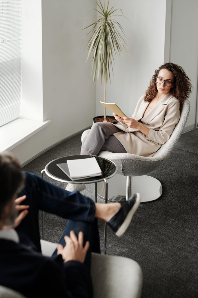 A woman with curly hair and glasses sits in a modern chair holding a notebook, engaged in conversation with a man whose back is to the camera. A small table with documents is between them, and a potted plant is in the background.