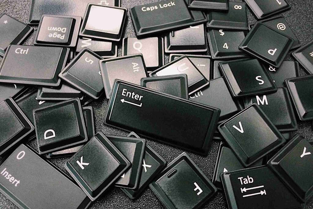 Loose black keyboard keys scattered on a dark surface. Various keys, including Enter, Caps Lock, Ctrl, Alt, and numbers, are visible. The arrangement is random, depicting disassembled keyboard parts.