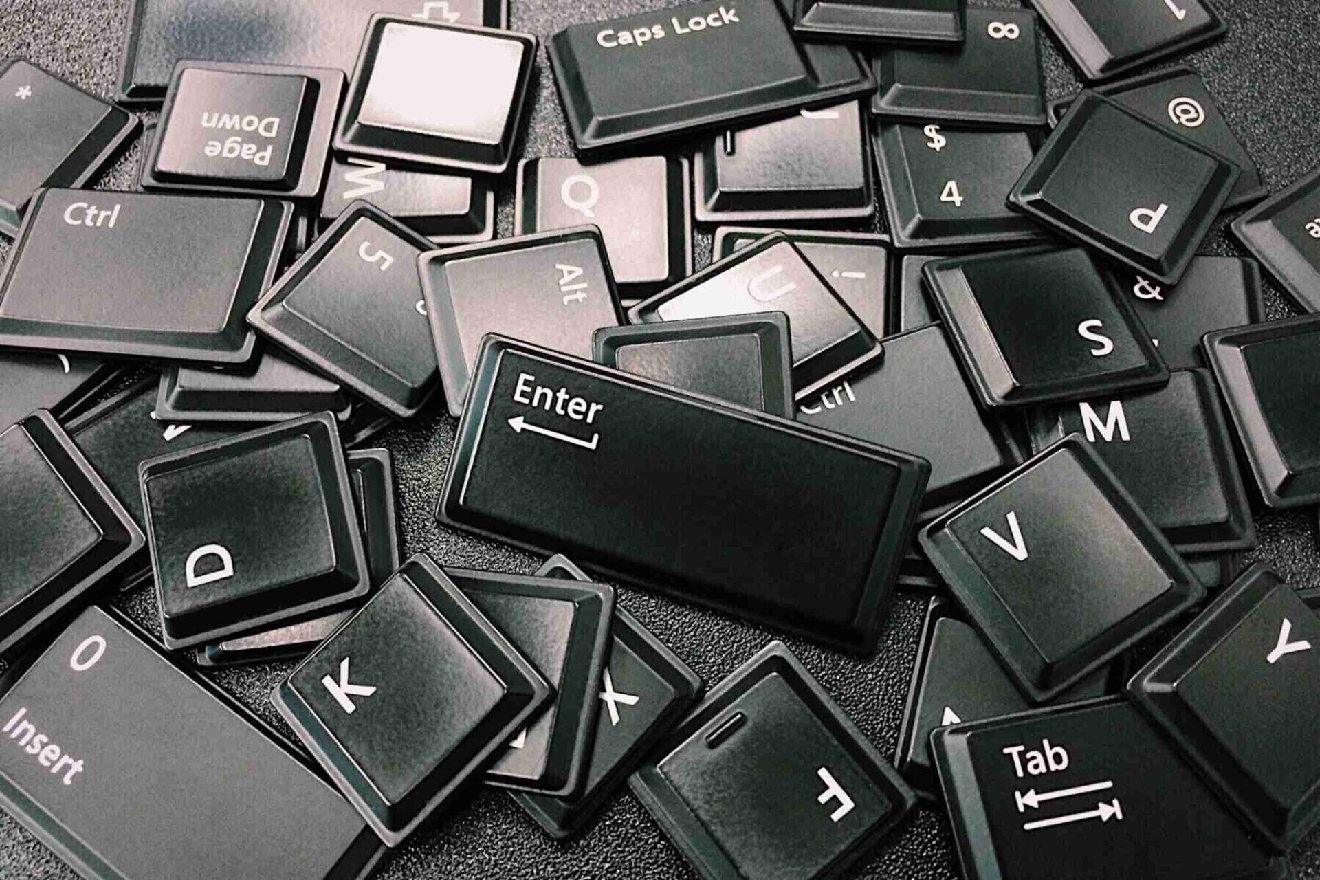 Loose black keyboard keys scattered on a dark surface. Various keys, including Enter, Caps Lock, Ctrl, Alt, and numbers, are visible. The arrangement is random, depicting disassembled keyboard parts.