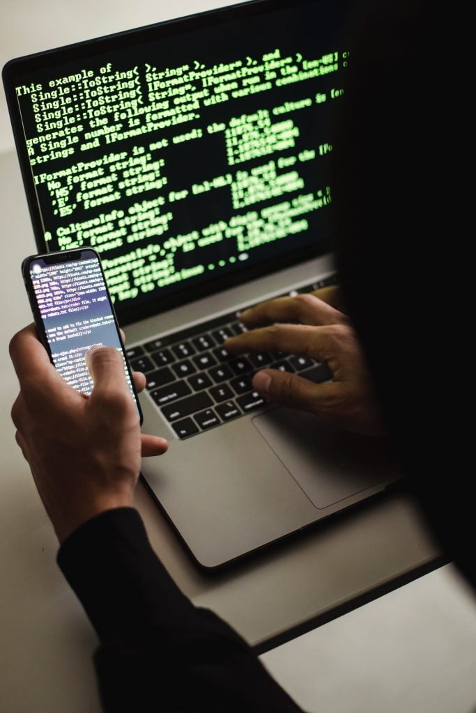 A person typing on a laptop with green code displayed on the screen. They are also holding a smartphone with similar code displayed on its screen. The image suggests multitasking or coding on multiple devices simultaneously.