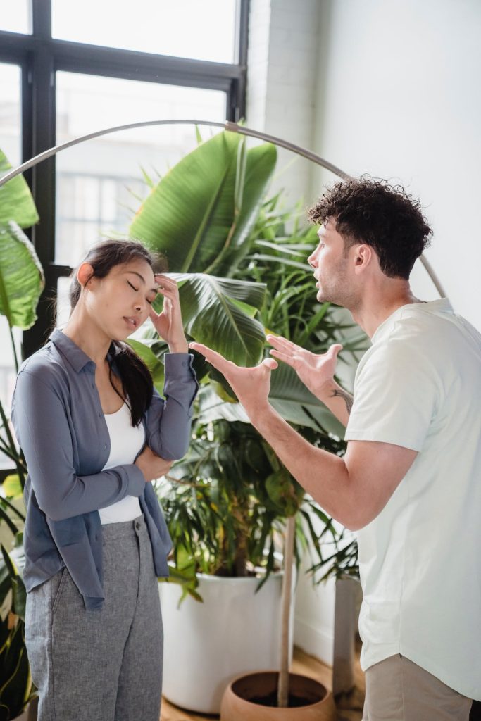A woman stands with her arms crossed and eyes closed, rubbing her forehead, appearing frustrated. A man, who is talking animatedly with hands raised, stands in front of her. They are indoors with large windows and potted plants in the background.