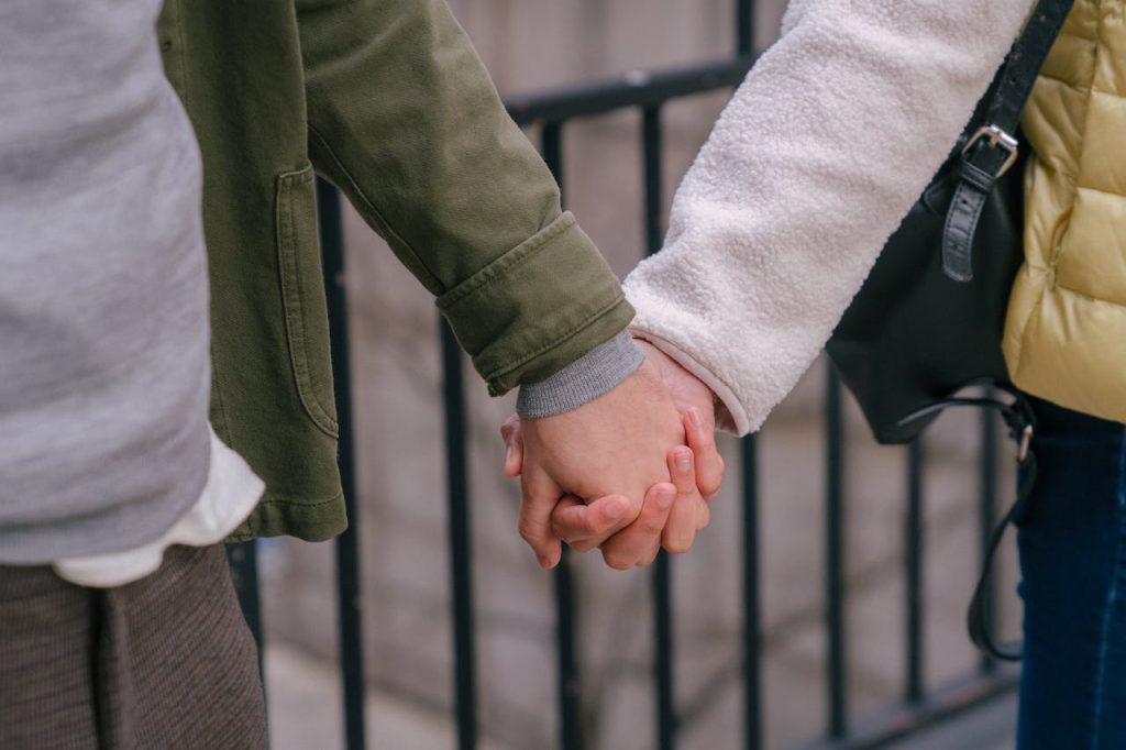 Two people holding hands, wearing winter clothing. The person on the left wears a green jacket, while the one on the right wears a yellow jacket. They are standing near a black fence. Only their hands and a portion of their torsos are visible.