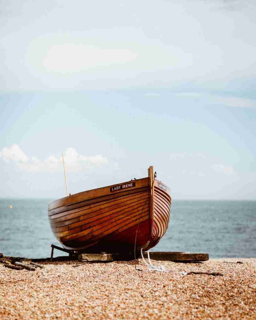 A small wooden boat named "Lady Irene" rests on a pebble beach with the sea and a partly cloudy sky in the background. The boat is slightly elevated on wooden supports, and its rich, polished wood contrasts with the light-colored pebbles.