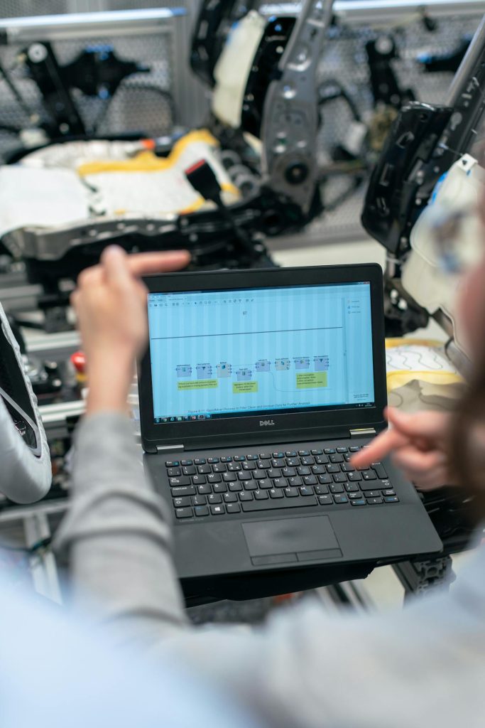 Person using a Dell laptop with a detailed chart on the screen, sitting among machinery parts in an industrial or manufacturing setting. The person's hands are visible, gesturing towards the chart, indicating a discussion or analysis in progress.