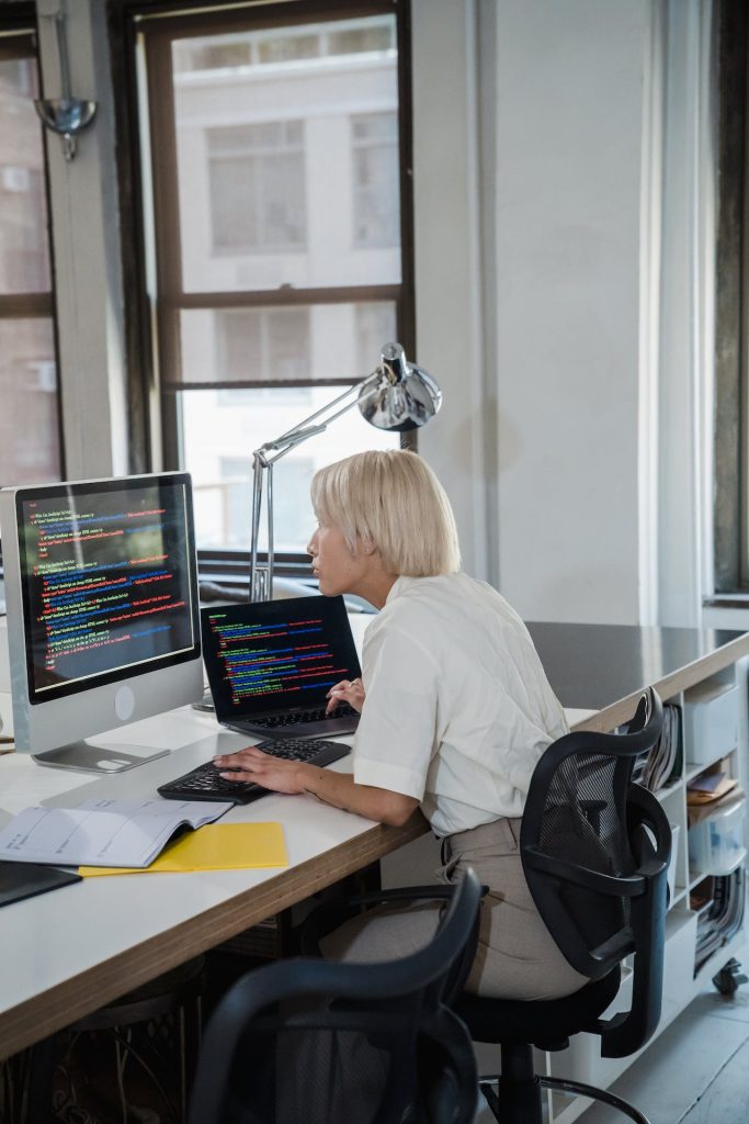A person with short blonde hair is seated at a desk in an office, working on a laptop and additional monitor displaying lines of code. They are focused on the screens, surrounded by office supplies, with large windows in the background letting in natural light.