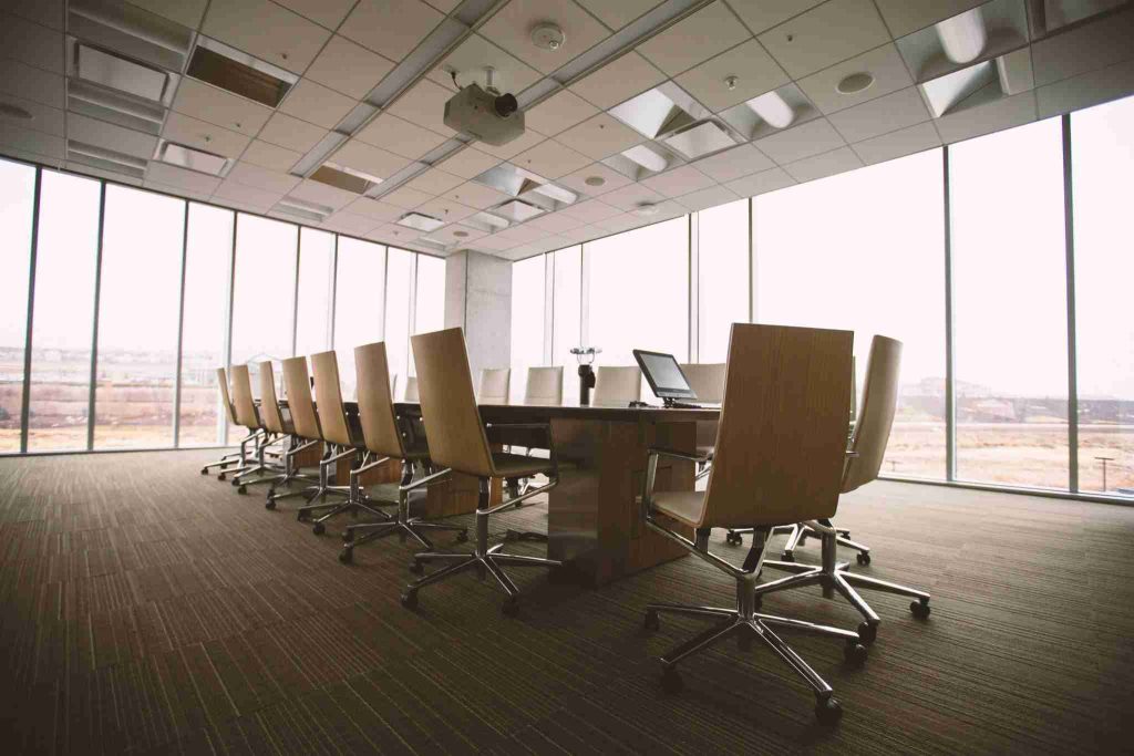 A large conference room with a long table surrounded by tan swivel chairs. The room, often used for commercial rent lease dispute mediations, has a high ceiling with recessed lighting and a projector. Floor-to-ceiling windows provide a view of an outdoor landscape. Laptops and office supplies are on the table.