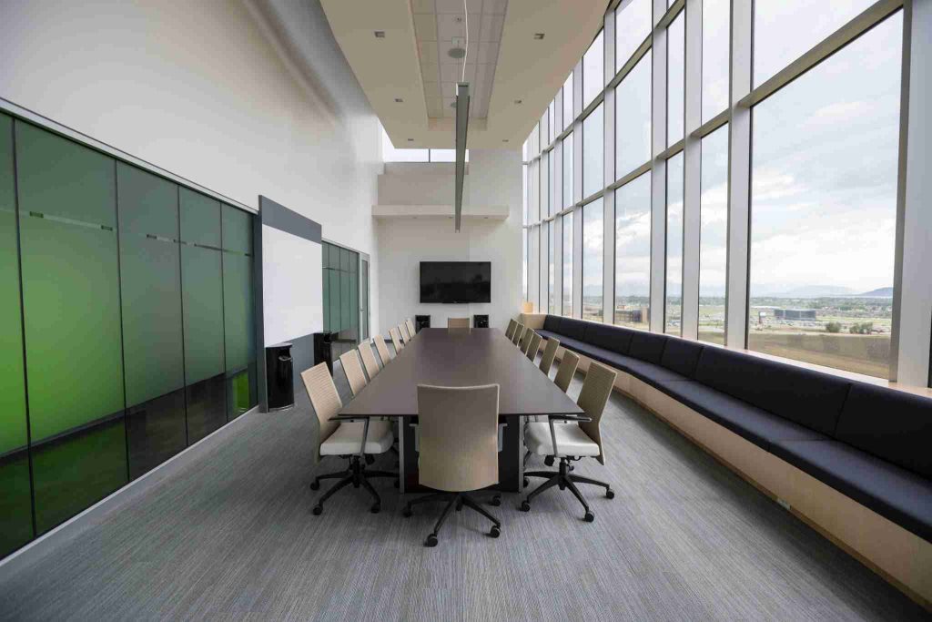 A modern conference room with a long rectangular table surrounded by swivel chairs, a large screen mounted on one wall, floor-to-ceiling windows providing natural light and views of the outdoors, and a strip of dark seating along the windows.