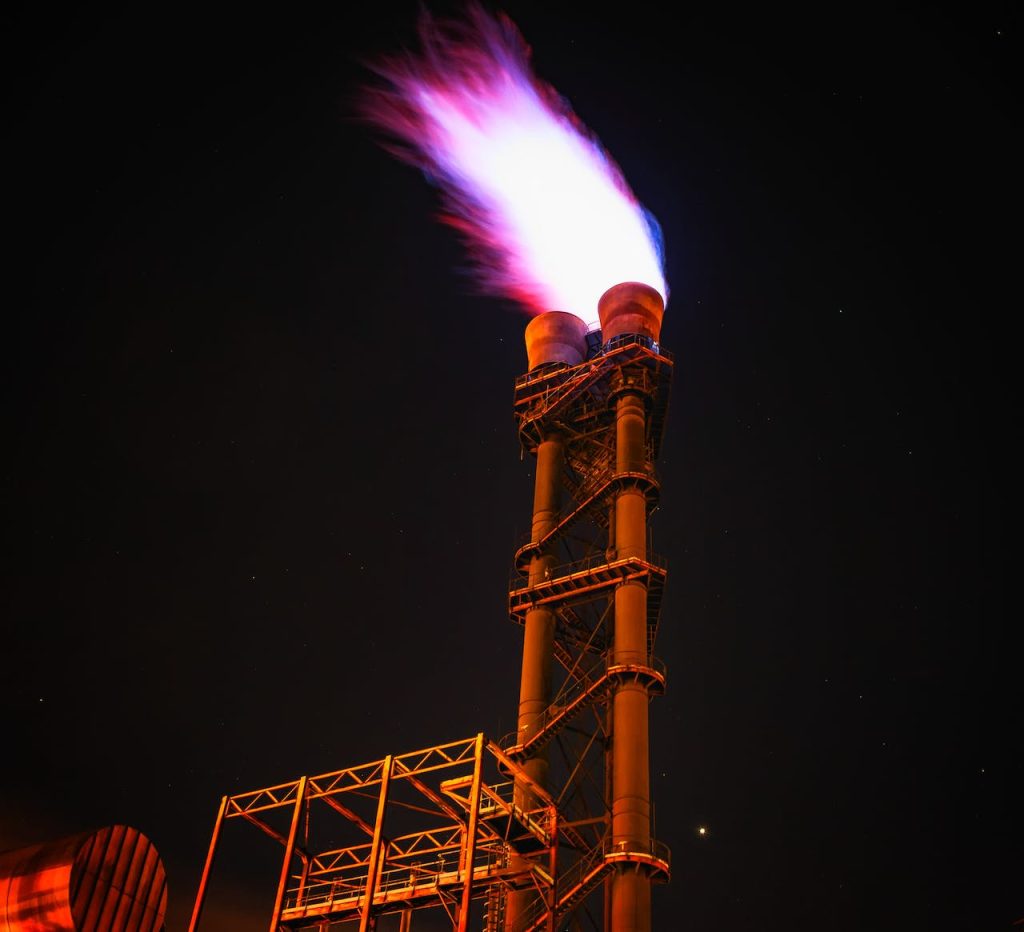 A tall industrial flare stack releases a bright, intense flame against a dark night sky. The structure is illuminated, revealing metallic frameworks and ladders, with scattered stars faintly visible in the background.