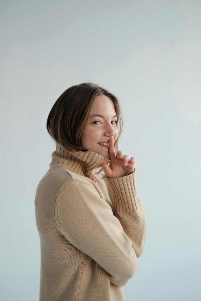 A woman with shoulder-length brown hair, wearing a beige turtleneck sweater, is standing against a light blue background. She is smiling and holding a finger to her lips in a "shh" gesture.