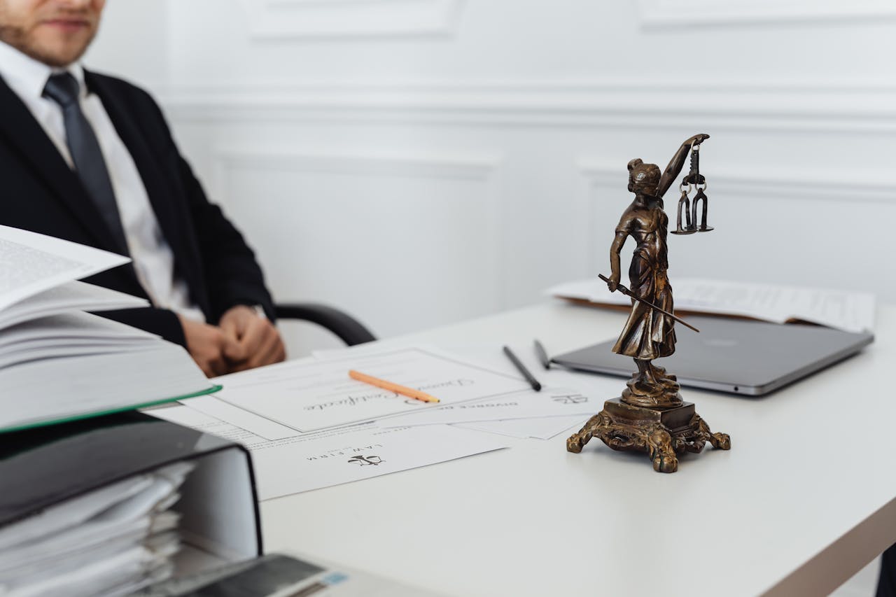 A man in a suit sits at a desk with various documents, a laptop, and a small bronze statue of Lady Justice prominently placed in the foreground. The focus is on the statue, with the man and background slightly blurred.