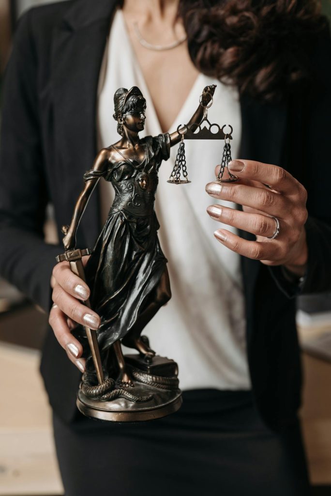 A woman in a white blouse and black blazer holds a bronze statue of Lady Justice, who is depicted wearing a blindfold and holding a balance scale and a sword. The woman's nails are painted silver, and she wears a silver ring.