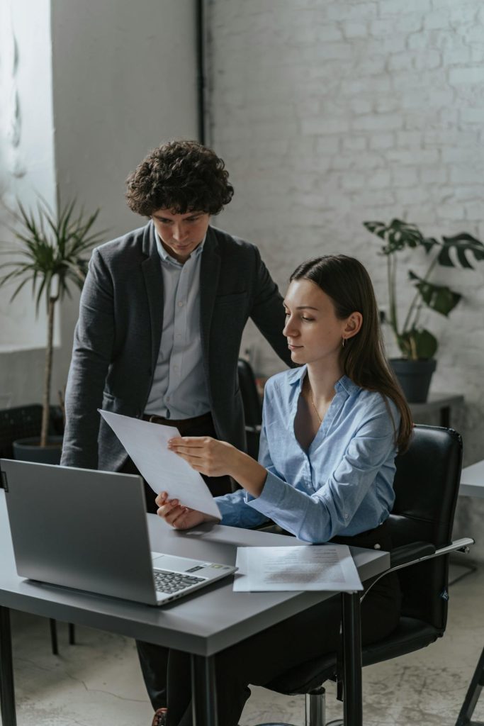 A woman sitting at a desk looks at a piece of paper while a man stands beside her, also looking at the paper. A laptop is open on the desk, and papers are scattered around. The setting appears to be an office with white brick walls and potted plants.