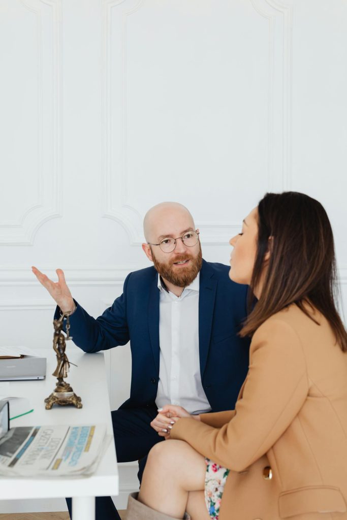 A man in a blue suit and glasses is sitting at a desk, gesturing with his hand, and talking to a woman with long dark hair in a tan blazer. They are in a white room with a white desk that has a small statue, a newspaper, and a pair of glasses on it.