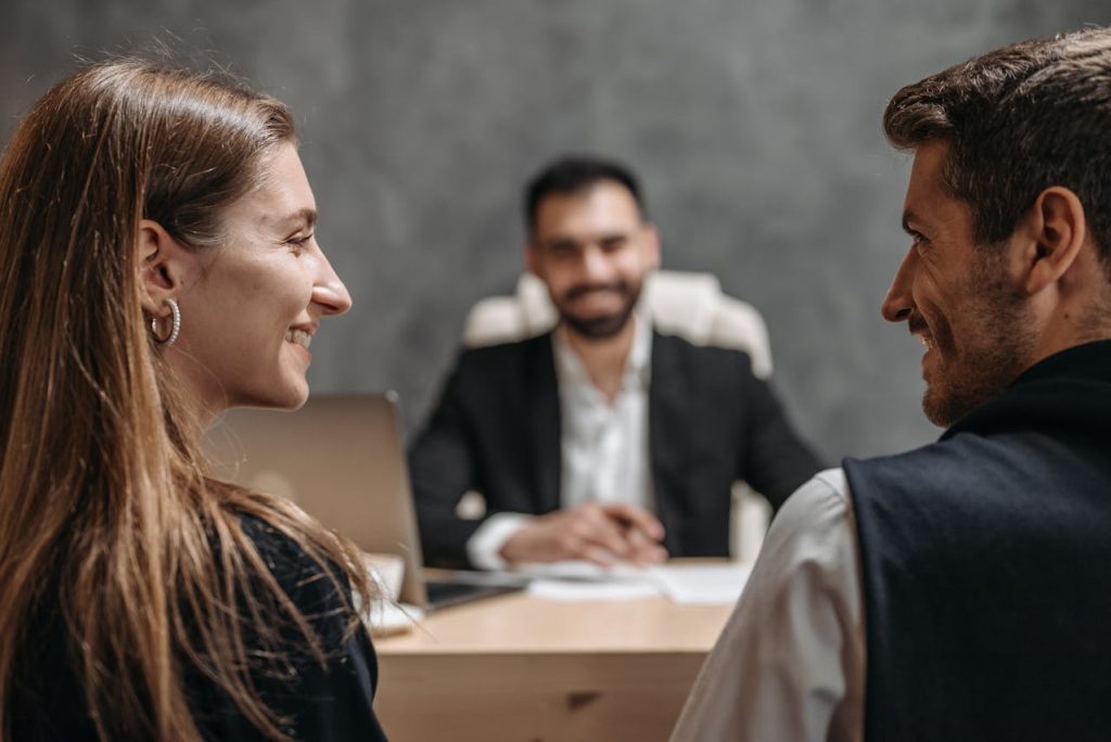 A man and woman sit across a desk from another man in a suit. The woman and man are smiling at each other, while the man in the suit, who is blurred in the background, appears to be facing them, seated, possibly for a formal discussion or meeting.