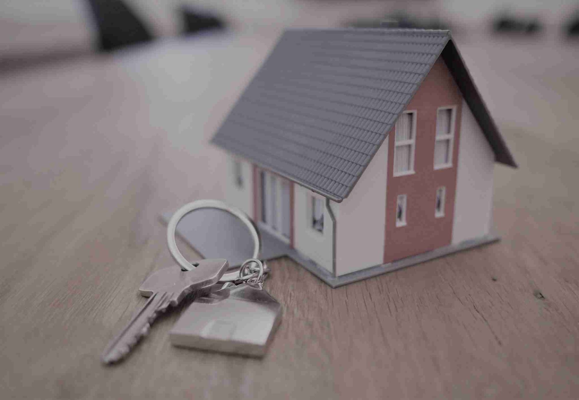 A small model house with a gray roof and white walls sits on a wooden surface. Beside the model house are a set of silver keys attached to a keychain, symbolizing real estate or home ownership.