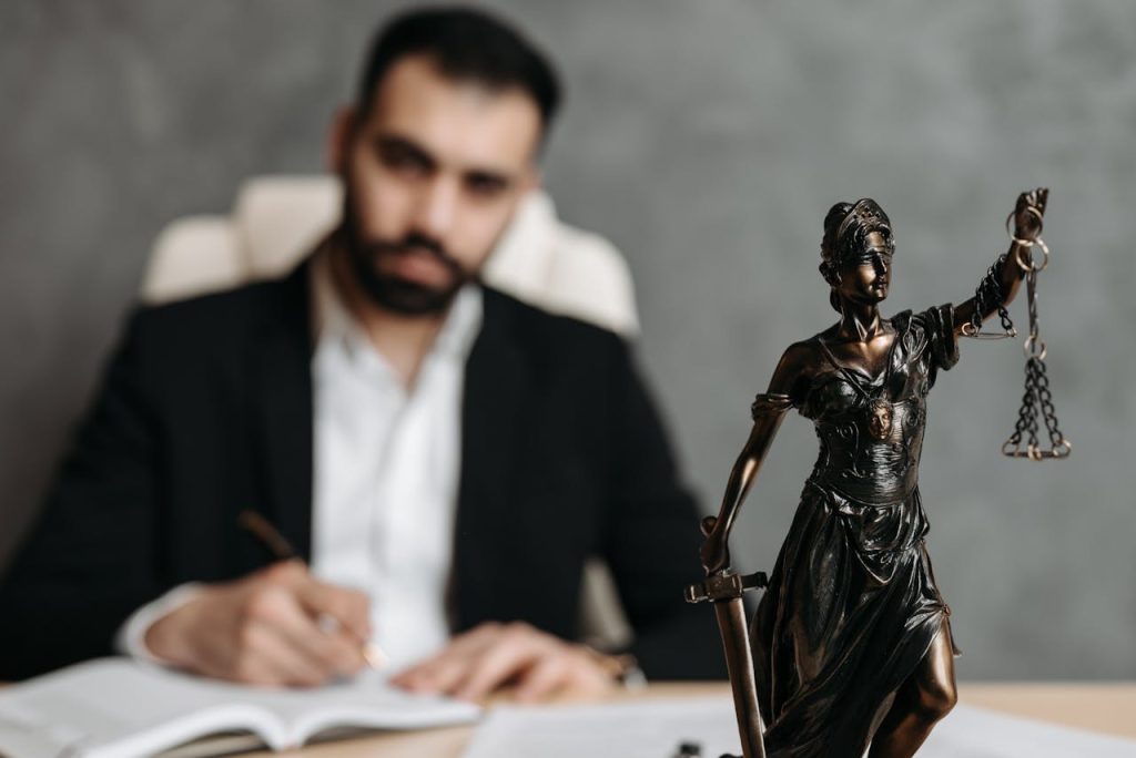 A man in a black suit sits at a desk, writing on a document. In the foreground, there is a bronze statue of Lady Justice holding scales and a sword. The background is blurred, emphasizing the statue.