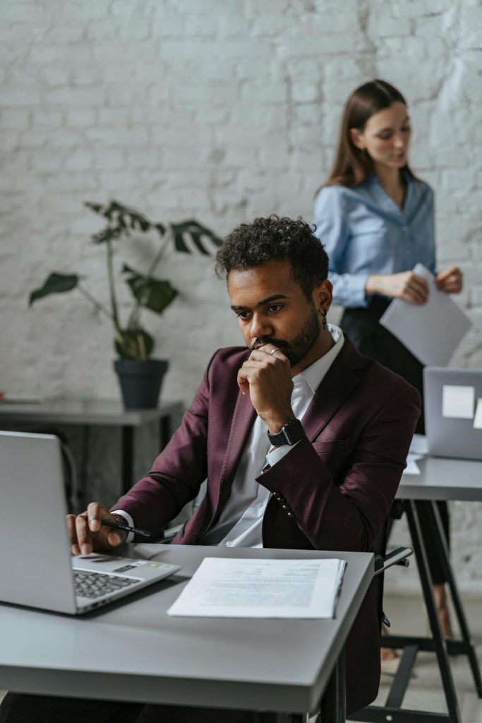 A man in a burgundy blazer sits at a desk, intently looking at his laptop with one hand on his chin. In the background, a woman in a blue shirt holds papers, standing near a brick wall with a potted plant. Both are in a modern office setting.