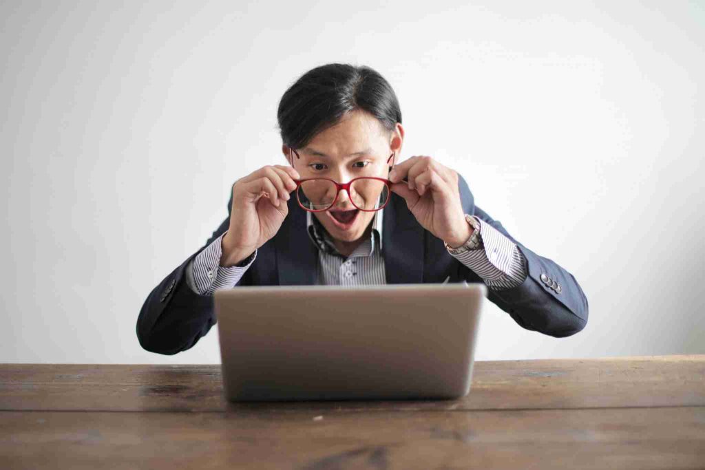 A man with glasses is sitting at a wooden table, looking surprised while peering at a laptop screen. He is lifting his glasses slightly off his face, eyes wide open, and the background is plain white.