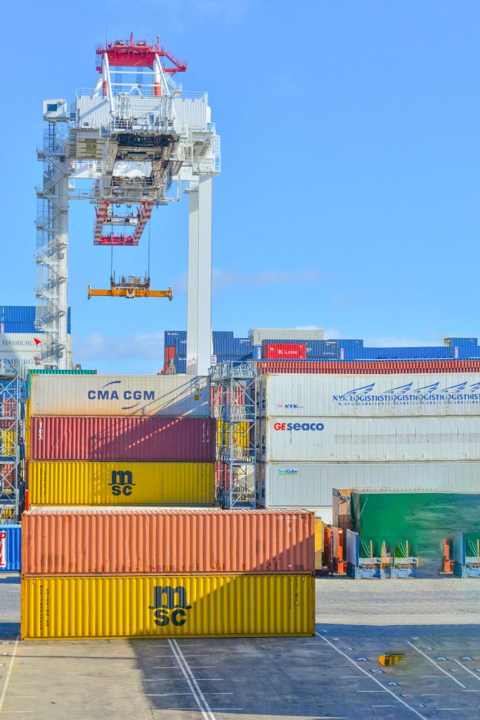 A seaport cargo yard showing multiple stacked shipping containers in varying colors including yellow, brown, and white. A large industrial crane in the background is lifting a container under a clear blue sky. Logos of various shipping companies are visible, emphasizing the importance of accuracy to prevent international arbitration errors.