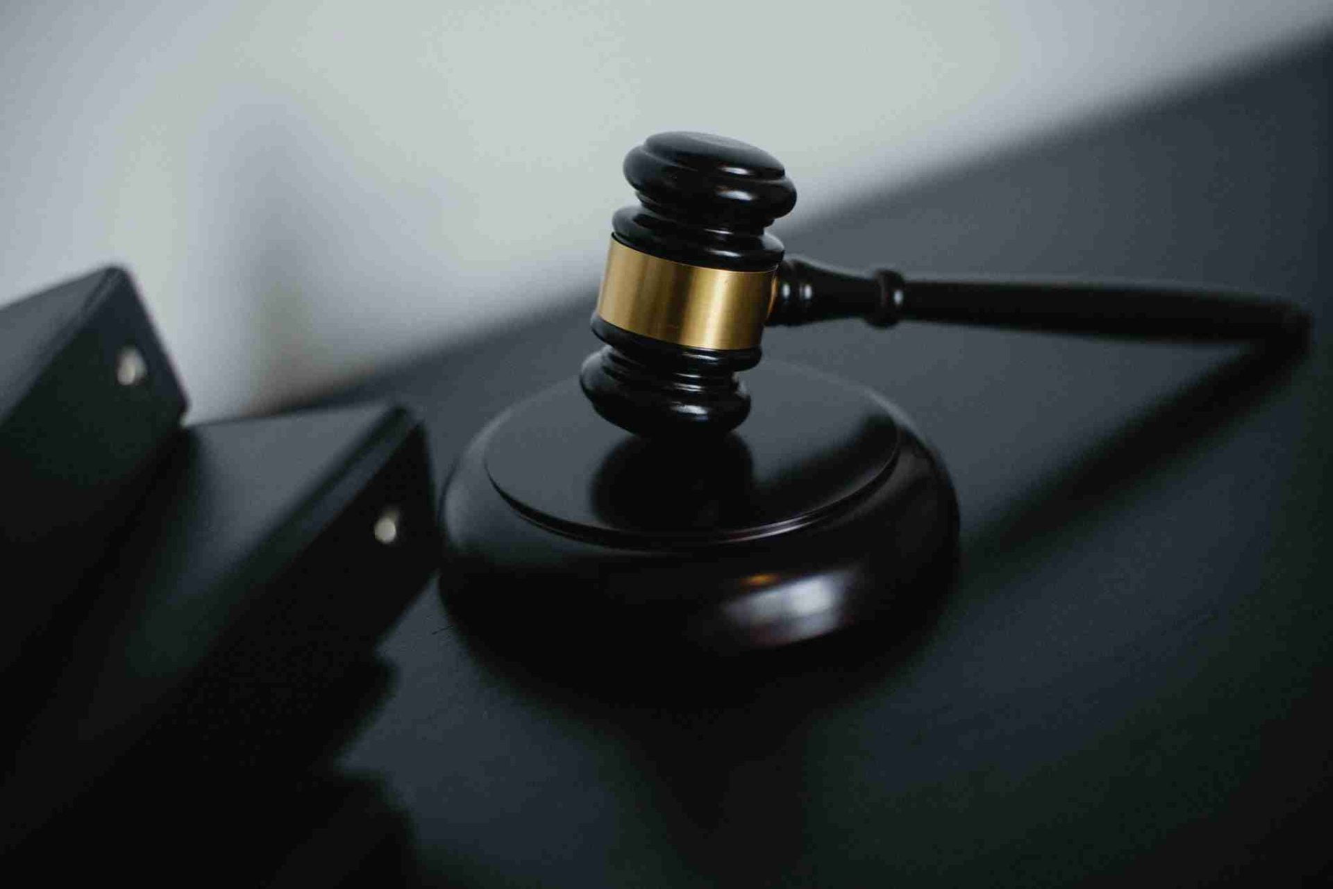 A close-up image of a wooden gavel with a brass band, placed on a sound block, typically used by judges in a courtroom setting. The gavel and sound block are situated on a dark surface, with two black binders visible in the background.