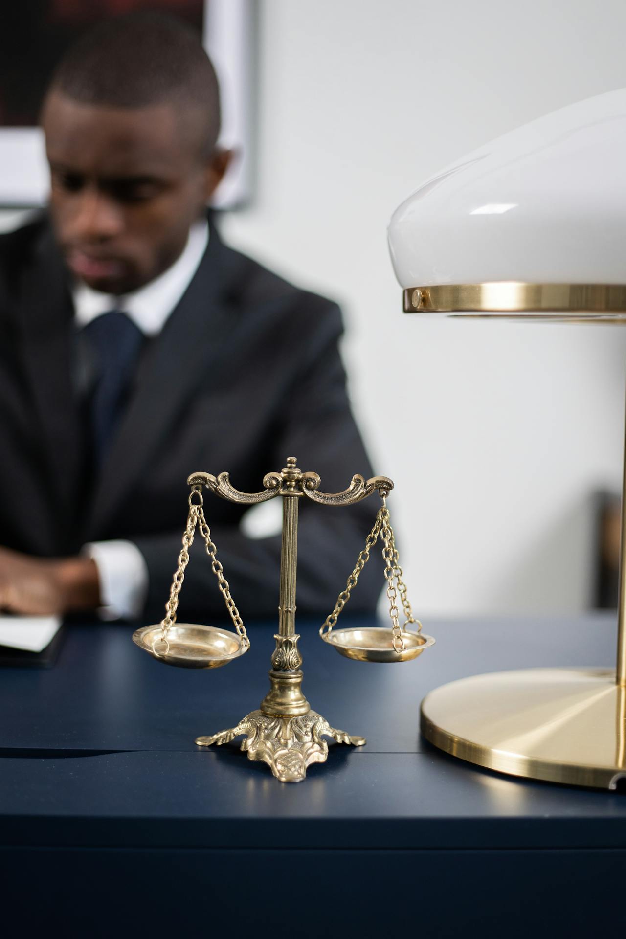 A small, golden balance scale sits on a dark blue desk. To the side is a modern white lamp with gold accents. In the background, a man in a dark suit works at the desk, out of focus, likely reviewing the limits to appeals of arbitral awards.