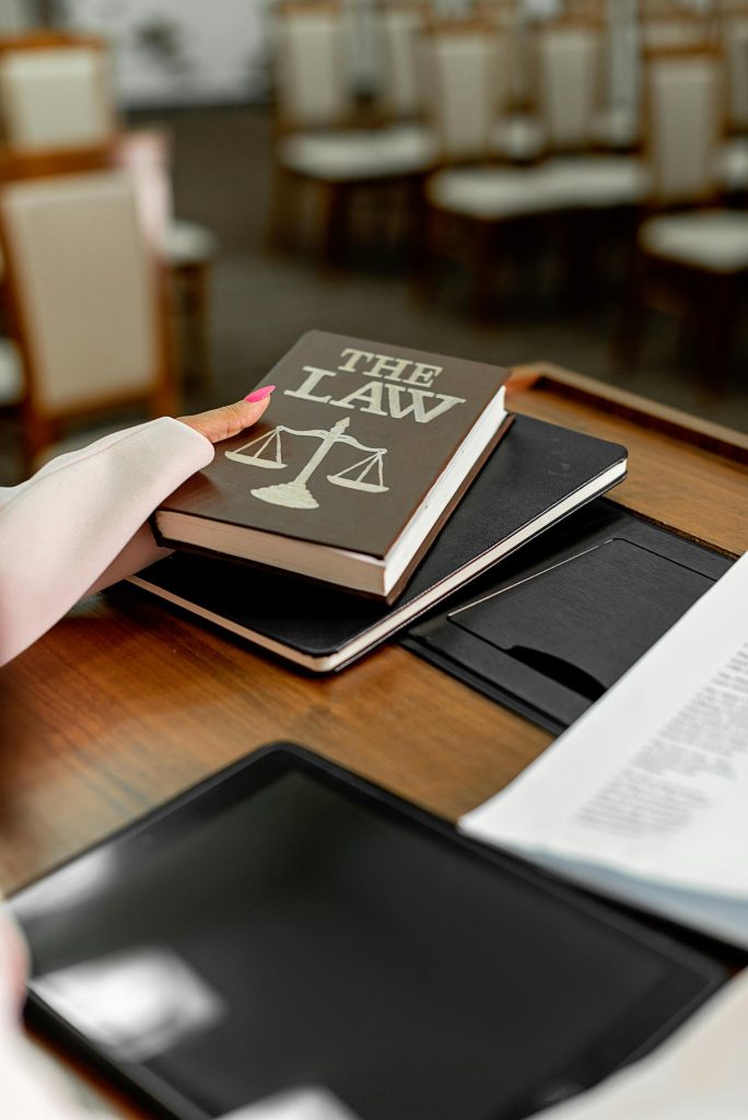 A person with pink nail polish holds a book titled "THE LAW," which features a scales of justice symbol on the cover. The book, discussing LIMITS TO APPEALS OF ARBITRAL AWARDS, rests on a closed laptop on a wooden table, next to a stack of papers and an open tablet. Rows of chairs are visible in the background.