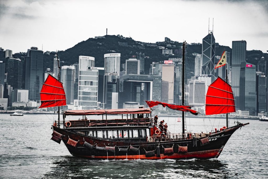 A traditional Chinese junk boat with vibrant red sails glides on the water in front of a modern city skyline featuring tall skyscrapers. The background includes a mountainous landscape under a cloudy sky, subtly hinting at the cost implications of challenging an arbitral award through the courts.