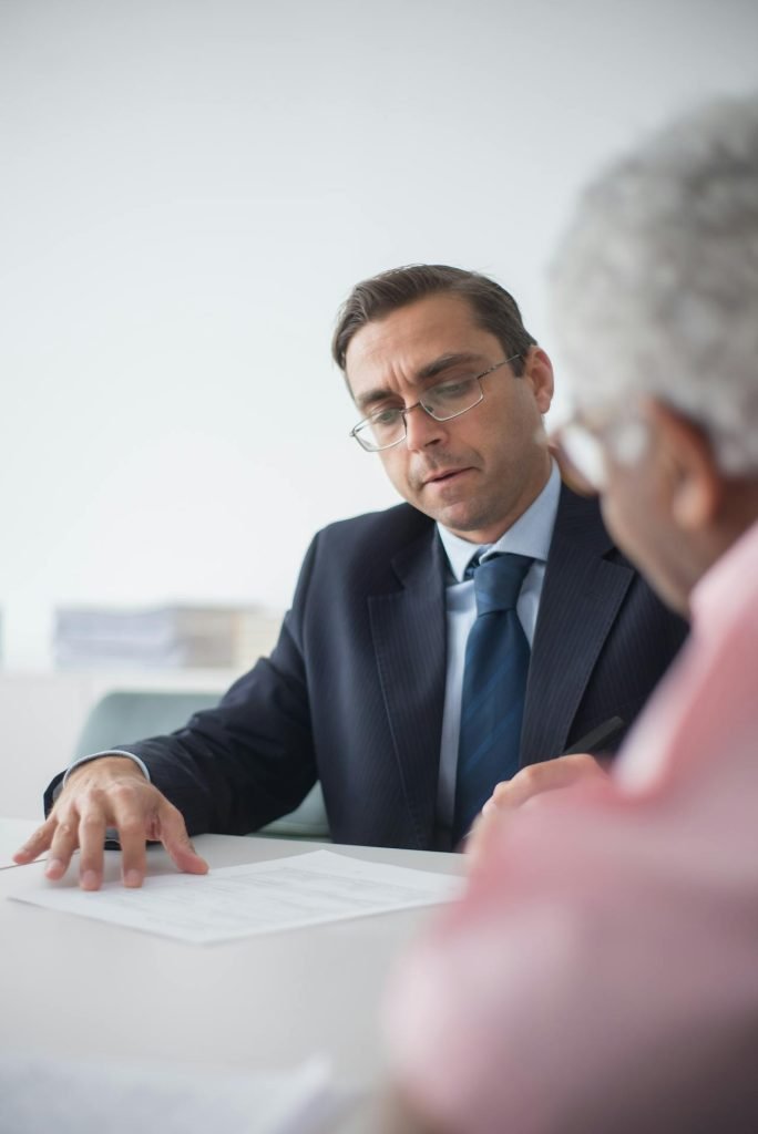 A man in a suit and glasses is seated at a table, attentively reviewing and discussing documents related to equitable compensation allowed for breach of arbitration clause by a third party with another person facing him, who has gray hair and is wearing a pink shirt. The background is softly lit, and office supplies are visible in the distance.