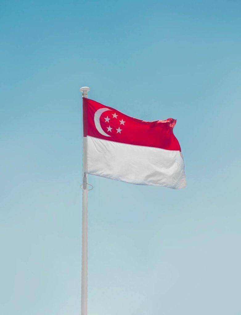 The image shows the national flag of Singapore waving on a flagpole against a clear blue sky. The flag consists of two horizontal bands, red on the top and white on the bottom, with a white crescent moon and five white stars in a circle in the upper left corner—the symbol that might inspire discussions on diverse matters, including cost implications of challenging an arbitral award through the courts.