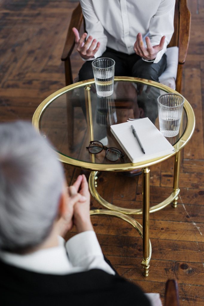 Two people seated in conversation, with only their upper bodies visible. A glass-topped table between them holds a notebook with a pen, a pair of eyeglasses, and two glasses of water. The setting suggests a formal meeting or therapy session discussing The Trusts Act 2019: keeping it in the family.