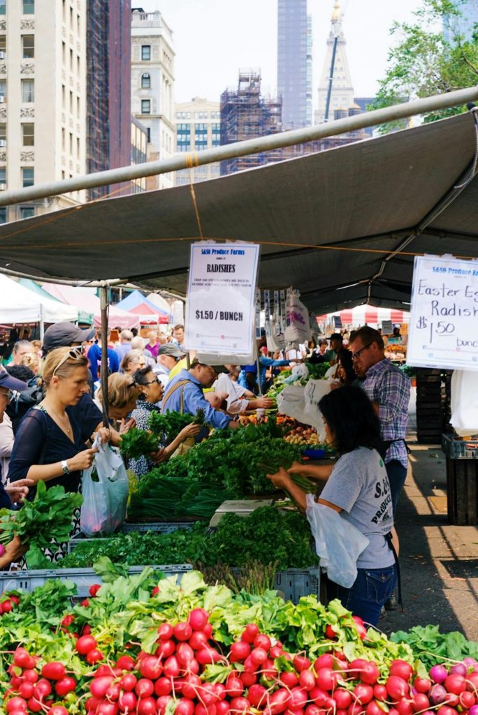 A bustling farmers' market with shoppers selecting fresh produce from vendor tables. The tables are filled with vibrant vegetables like radishes, greens, and carrots. A canopy provides shade, and the city's tall buildings are visible in the background. Important changes to the Fair Trading Act 1986 ensure fair treatment for all shoppers.