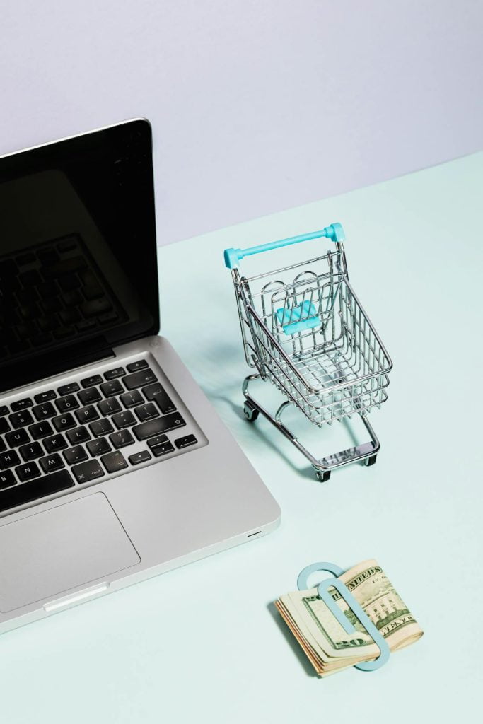 A laptop sits on a light blue surface next to a small shopping cart model and a stack of US dollar bills secured with a blue money clip, highlighting the importance of avoiding unfair contract terms in online shopping or e-commerce scenarios.