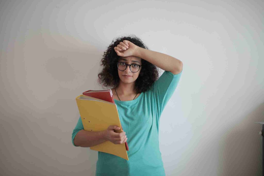 A woman with curly hair and glasses, dressed in a turquoise top, holds folders against her chest with one hand while resting her other arm on her forehead. She stands in front of a plain off-white wall, appearing tired or stressed.