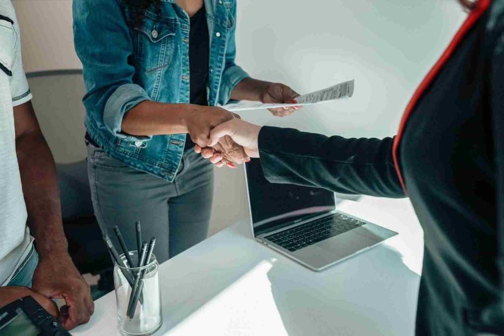 Two people shake hands over a desk with a laptop and a cup holding pens. One person is holding a piece of paper. Both individuals are wearing blazers, and part of a third person is partially visible to the left. The professional setting suggests ongoing discussions about the Fair Trading Amendment Bill at the New Zealand Dispute Resolution Centre.