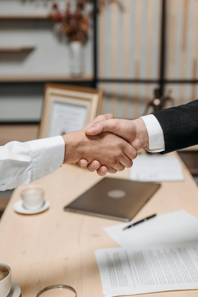 Two individuals are shaking hands over a desk. The desk has a laptop, a cup of coffee, a pen, a pair of glasses, and documents. One person is wearing a white shirt, and the other is in a black sleeve, possibly indicating a formal agreement or business deal.