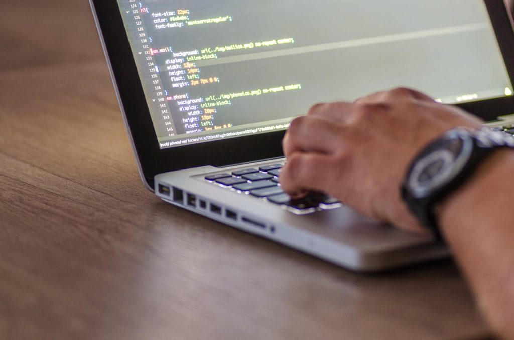 A person is typing on a laptop keyboard. The laptop screen displays lines of code in a text editor, perhaps analyzing a recent legal development like the UK Supreme Court returning to the orthodox liquidated damages approach. The person's left hand and wristwatch are visible. The background consists of a wooden surface.