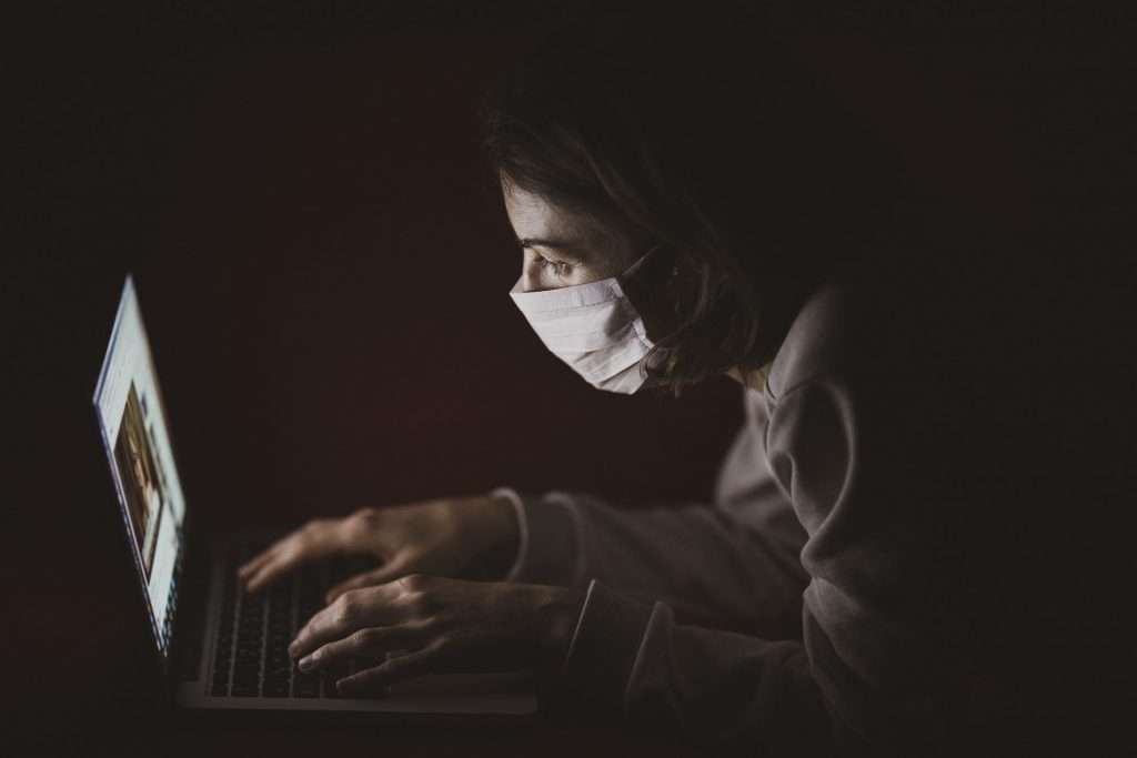 A person wearing a face mask is sitting in a dark room, illuminated by their laptop screen. Their hands are typing on the keyboard, perhaps drafting contracts or seeking insight from overseas courts during the pandemics lockdowns.