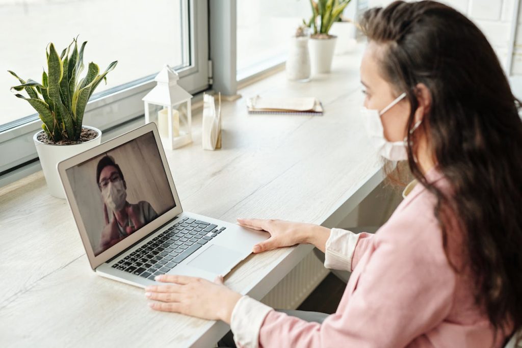 A woman wearing a mask is sitting at a desk, participating in a video call on her laptop during one of many pandemics lockdowns. The person on the screen is also masked. The desk features potted plants, a notebook, and small decor items, all set in a brightly lit room.