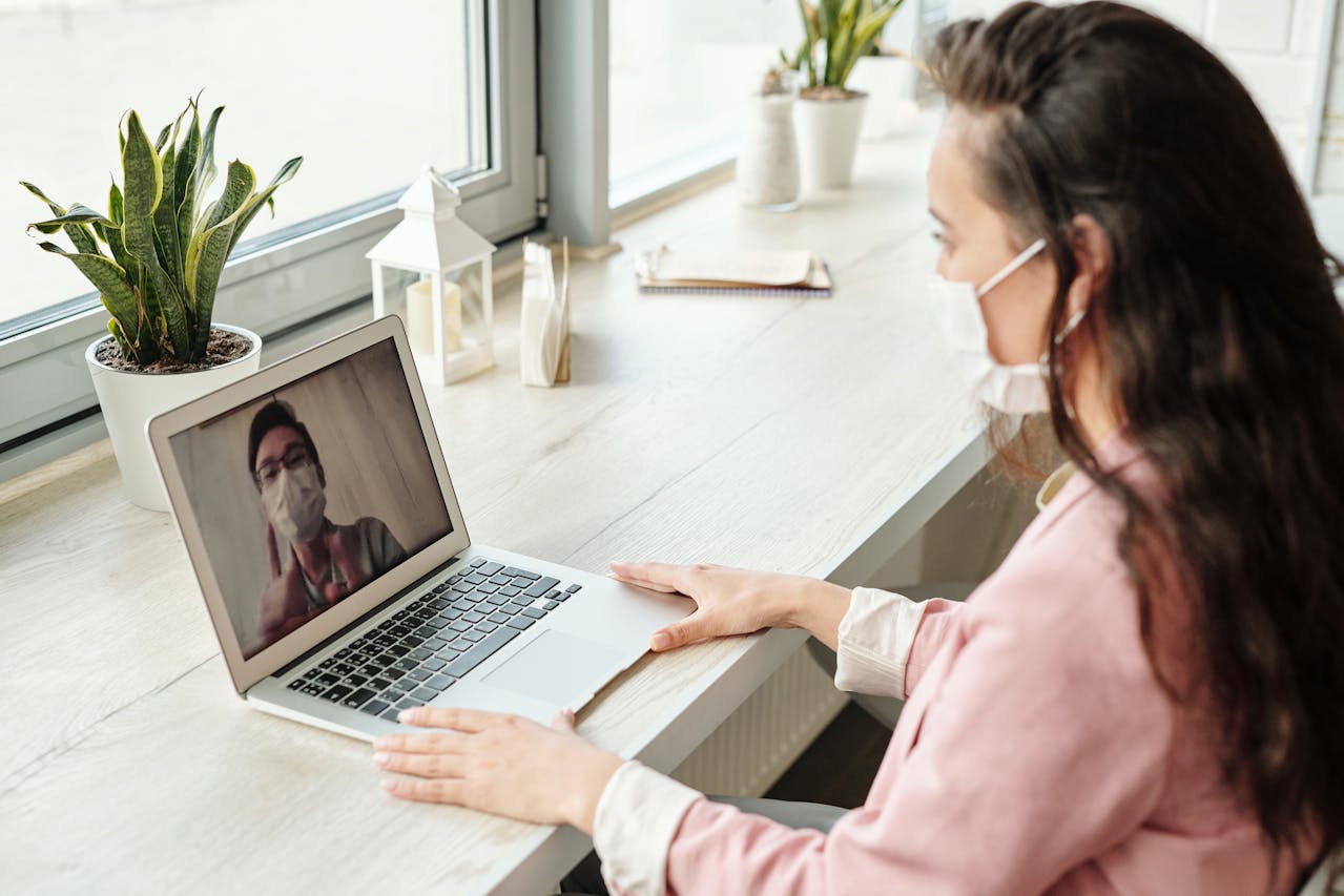 A woman wearing a mask is sitting at a desk, participating in a video call on her laptop during one of many pandemics lockdowns. The person on the screen is also masked. The desk features potted plants, a notebook, and small decor items, all set in a brightly lit room.