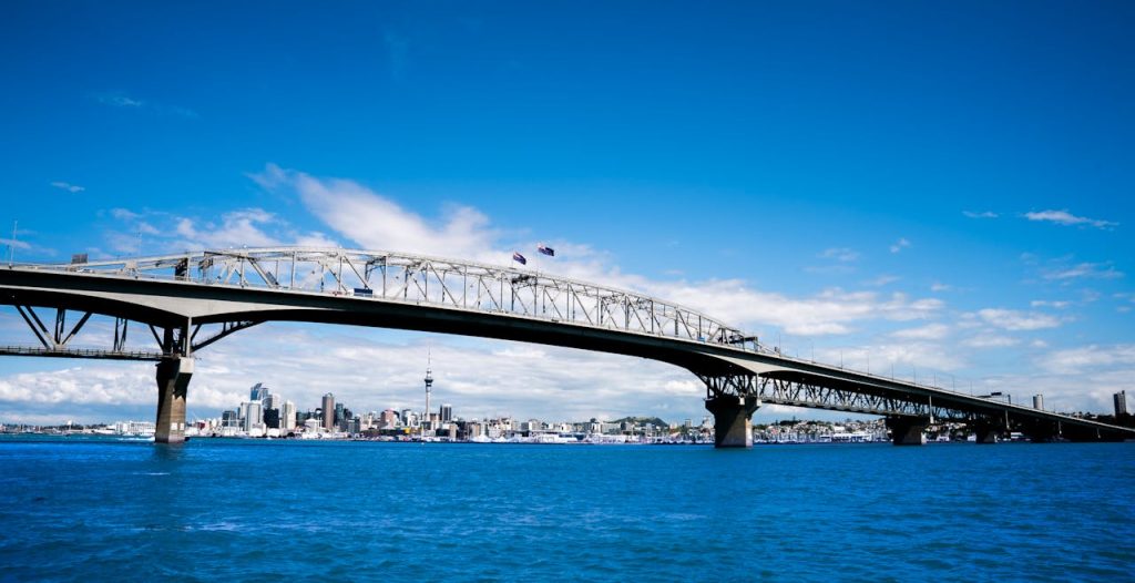 A large steel bridge spans a deep blue body of water on a bright, sunny day. The skyline of a city with tall skyscrapers and a prominent tower is visible in the background under a clear blue sky with some scattered clouds, reminiscent of bustling days before pandemics reshaped our routines.