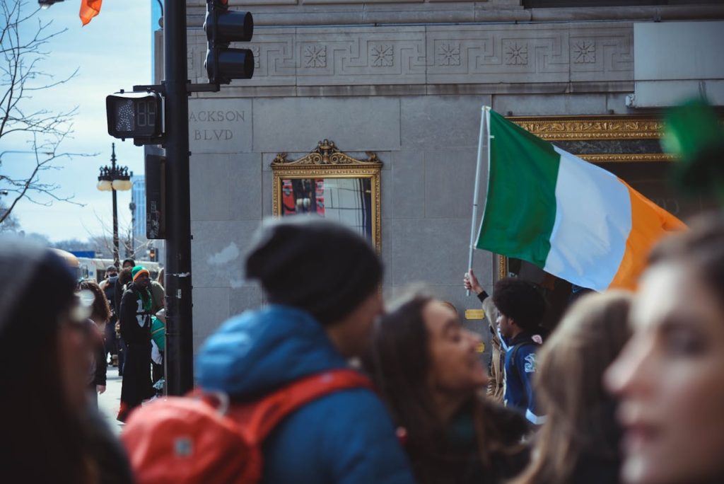 People are gathered on a city street for a parade. A person holds up a large Irish flag, which is prominently displayed. There is a traffic light and a street sign saying "Jackson Blvd" on the corner of a building in the background, reminiscent of times before pandemics lockdowns transformed public celebrations.