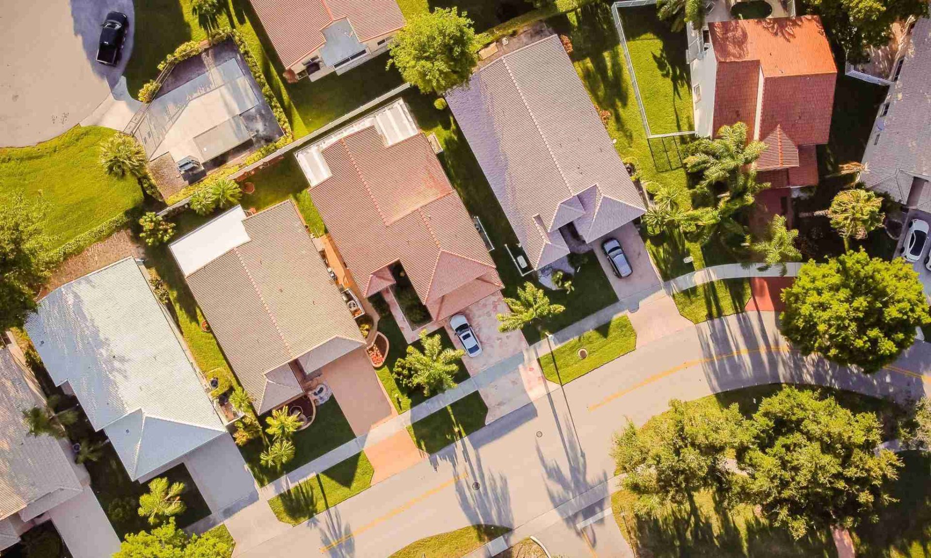 Aerial view of a suburban neighborhood featuring houses with yards, driveways, and cars parked on paved driveways. The street is lined with tall trees casting long shadows, and the homes have varied roof colors and styles, with greenery surrounding them.