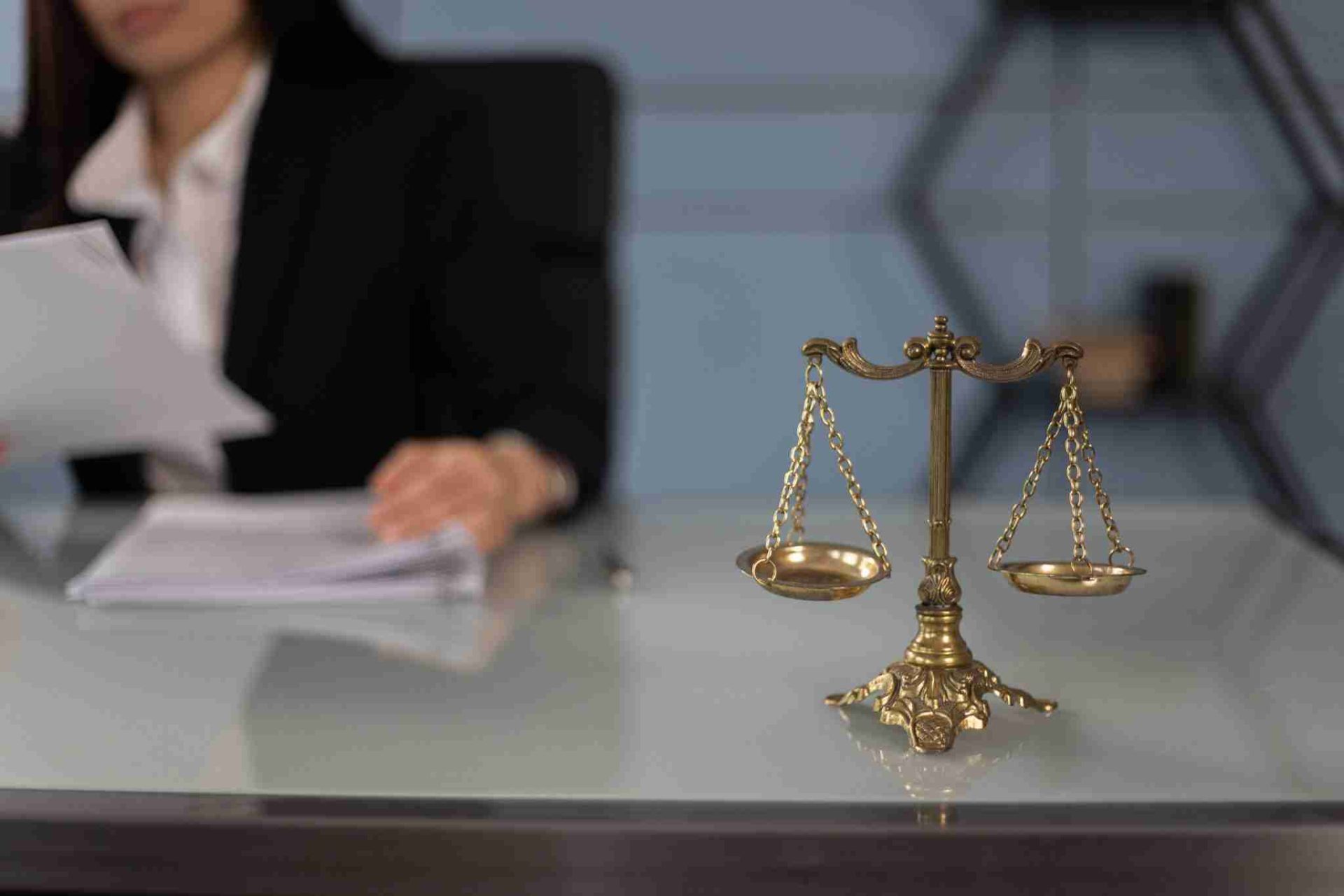 A brass scale of justice sits prominently on a glass desk, symbolizing law and fairness. In the background, a person in a suit, partially out of focus, examines documents, indicating that even in this seemingly straightforward setting, the court sees through layers of lipstick and linguistic camouflage.