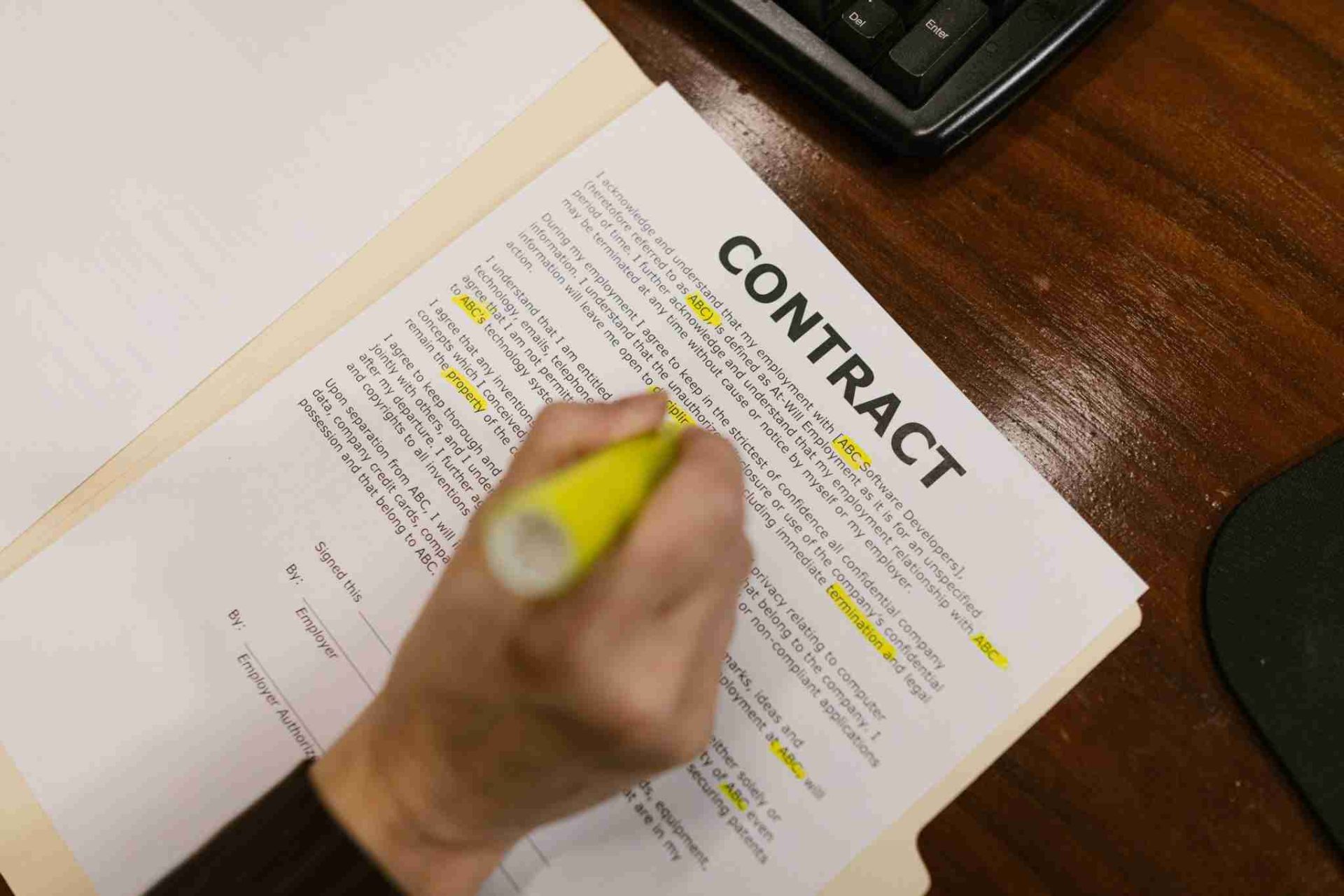 A person is highlighting text on a printed contract document with a yellow marker. The document, which discusses the UK Supreme Court's return to an orthodox liquidated damages approach, is placed on a wooden desk next to a keyboard and mouse. Various sections and terms have some text already highlighted.